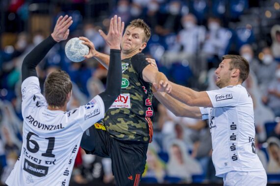 Ein Handballspieler in einem schwarz-grünen Trikot springt, um den Ball zu schießen, während zwei Verteidiger in weißen Trikots versuchen, ihn während eines Spiels zu blockieren, während im Hintergrund maskierte Zuschauer zu sehen sind.