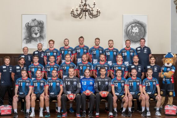 Eine Männerhandballmannschaft posiert in der Halle in Uniformen, flankiert von Trainern und einem Löwenmaskottchen. Die Gruppe ist in drei Reihen unter Kronleuchtern angeordnet, hinter ihnen hängen große gerahmte Löwenskizzen an der Wand.