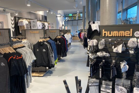 Ein modernes Sportbekleidungsgeschäft mit Reihen von Kleiderständern mit Trikots, Jacken und Hemden sowie Accessoires wie Schlüsselbänder und Schlüsselanhänger. Das Hummel-Branding ist auf mehreren Schildern zu sehen.