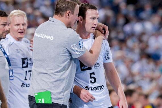 Ein Trainer ermutigt einen Handballspieler, der ein weißes Trikot mit der Nummer 5 trägt, während eines Spiels, während andere Mannschaftsmitglieder in der Nähe stehen und eine unscharfe Menschenmenge im Hintergrund zu sehen ist.
