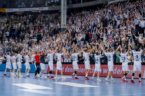 Eine Handballmannschaft in weißen Trikots steht in einer Reihe auf einem Hallenplatz, hält sich an den Händen und hebt sie in Richtung einer jubelnden, dicht gedrängten Menge auf der Tribüne. Die Halle ist hell erleuchtet und die Atmosphäre scheint feierlich.