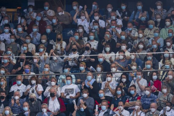 Eine große Menschenmenge, von denen viele Gesichtsmasken tragen, sitzt dicht beieinander in stadionähnlichen Sitzreihen, einige klatschen oder heben die Arme, was darauf hindeutet, dass sie ein Ereignis oder ein Spiel verfolgen.
