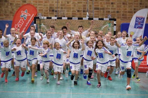 Eine große Gruppe aufgeregter Kinder in passenden weißen Sportuniformen läuft in einer Sporthalle auf die Kamera zu, lächelt und hebt die Arme. Im Hintergrund sind ein Handballnetz und Fahnen zu sehen.
