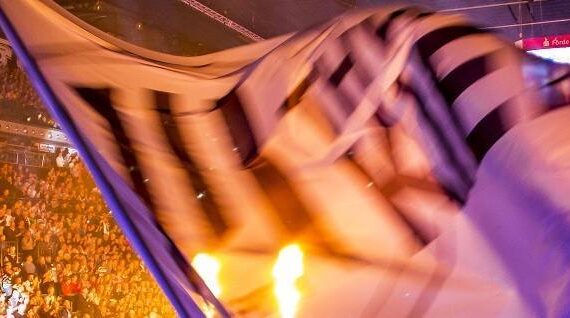 Eine große schwarz-weiße Fahne weht im Vordergrund einer Hallenveranstaltung, die Tribünen sind mit einer jubelnden Menge gefüllt, und die Szene ist hell erleuchtet.