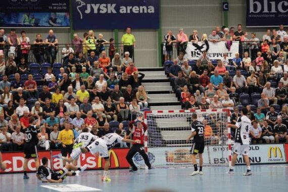 Bei einem Handballspiel in einer Halle sind die Spieler in der Nähe des Tores in Aktion, wobei ein Spieler zu Boden fällt. Die Tribünen sind mit Zuschauern gefüllt, die das Spiel aufmerksam verfolgen.