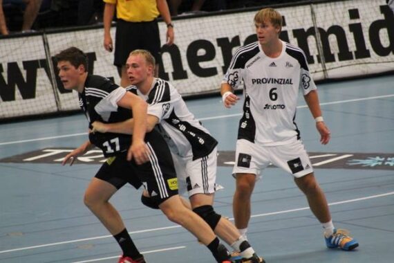 Drei männliche Handballspieler in Aktion auf einem Hallenplatz; zwei Spieler in Schwarz werden während eines Spiels von einem Spieler in Weiß eng markiert. Im Hintergrund sind Zuschauer und Beschilderung zu sehen.