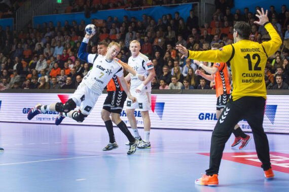Ein Handballspieler in einem weißen Trikot springt auf, um den Ball zu schießen, während orangefarbene Verteidiger versuchen, ihn zu blockieren; der Torwart in Gelb und Schwarz bereitet sich darauf vor, den Schuss zu stoppen. Eine Menschenmenge schaut von der Tribüne aus zu.