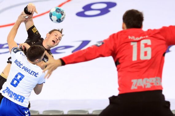 Ein Handballspieler in Schwarz versucht zu schießen, während er von einem Spieler in Weiß und Blau verteidigt wird, während sich ein Torwart in einem roten Trikot mit der Nummer 16 darauf vorbereitet, den Schuss zu blocken.