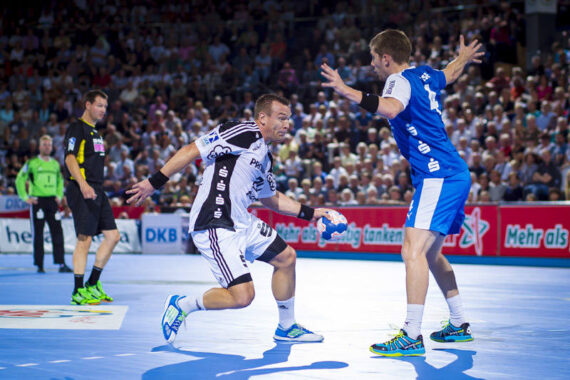 Ein Handballspieler in einem schwarz-weißen Trikot dribbelt den Ball, während er von einem Spieler in Blau verteidigt wird, auf einem Hallenplatz mit einem Schiedsrichter und einer Menschenmenge im Hintergrund.