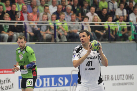 Ein Handballspieler in einem weißen Trikot hält den Ball und bereitet sich auf einen Spielzug vor, während ein Gegner in einem grünen Trikot zuschaut. Das Spiel findet in einer Halle statt, während die Zuschauer von der Tribüne im Hintergrund zusehen.