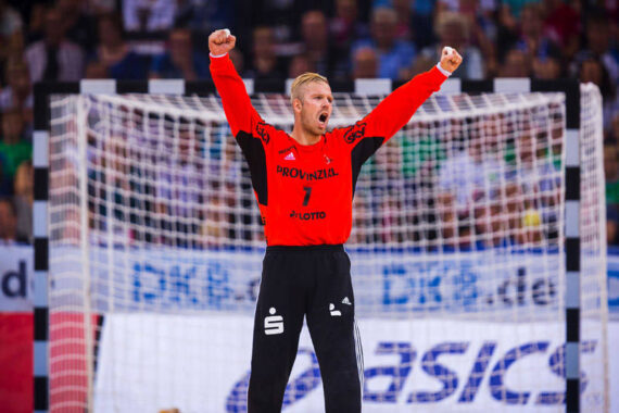 Ein männlicher Handballtorwart in einem leuchtend roten Trikot mit erhobenen Armen und offenem Mund, der während eines Spiels feiert. Der Hintergrund zeigt eine verschwommene Menge und einen Teil des Tornetzes.