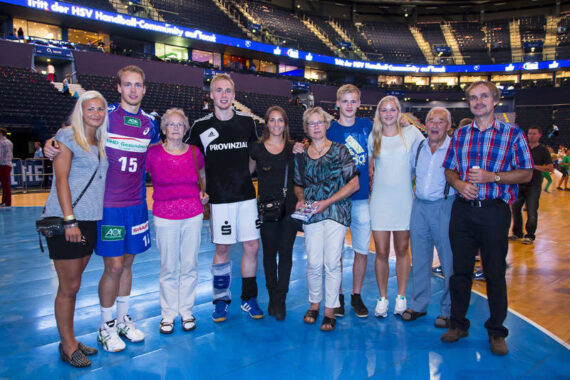 Eine Gruppe von zehn Personen, darunter Sportler in Sportuniformen und andere in Freizeit- oder Abendgarderobe, stehen zusammen auf einem Sportplatz und posieren und lächeln für ein Foto in einer großen Arena mit leeren Sitzen hinter ihnen.