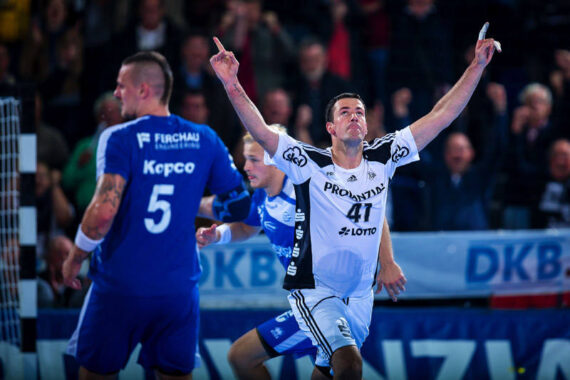 Ein Handballspieler in einem weiß-schwarzen Trikot reißt beide Arme in die Höhe und jubelt, während Spieler in blauen Trikots in der Nähe des Spielfelds stehen und die Menge im Hintergrund jubelt.