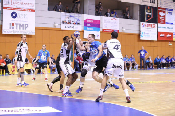 Ein Handballspiel in Aktion mit Spielern in Schwarz und Weiß, die gegen einen Spieler in Blau verteidigen, der springt und den Ball hält, während Mannschaftskameraden und Zuschauer vom Spielfeldrand aus zusehen.