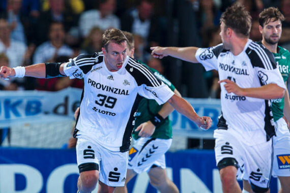 Zwei männliche Handballspieler in weißen Trikots, einer mit der Nummer 39, feiern auf einem Hallenplatz. Im Hintergrund sind ein Spieler in einem grünen Trikot und Zuschauer zu sehen.