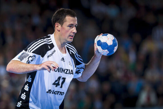 Ein männlicher Handballspieler in einem weiß-schwarzen Trikot mit der Nummer 41 bereitet sich während eines Spiels auf den Wurf eines blau-weißen Balls vor, während im Hintergrund eine unscharfe Menschenmenge zu sehen ist.