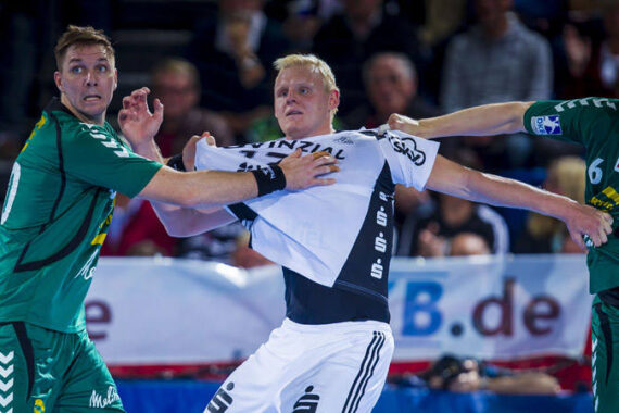 Zwei Handballspieler in grünen Trikots greifen und ziehen am Trikot eines gegnerischen Spielers in einer weiß-schwarzen Uniform während eines intensiven Spiels. Die Szene fängt Körperkontakt und Wettbewerb ein.