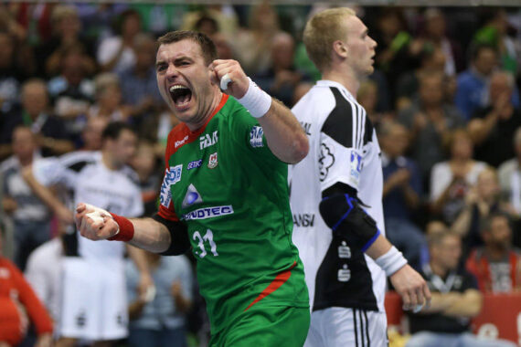 Ein männlicher Handballspieler in einem grünen Trikot jubelt leidenschaftlich, ballt die Fäuste und schreit, während ein anderer Spieler in einem weiß-schwarzen Trikot in der Nähe steht. Im Hintergrund ist eine unscharfe Menschenmenge zu sehen, die das Spiel verfolgt.