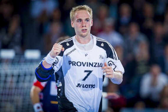 Ein männlicher Handballspieler, der ein weiß-schwarzes Trikot mit der Nummer 7 und dem Wort Provinzial trägt, jubelt während eines Spiels mit einer geballten Faust. Der Hintergrund zeigt eine unscharfe Menge und einen Teil des Tornetzes.