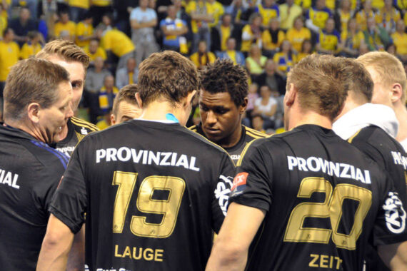 Eine Gruppe von männlichen Handballspielern in schwarzen Trikots drängt sich während eines Spiels zusammen. Auf den Trikots stehen die Namen Lauge und Zeit mit den Nummern 19 und 20. Zuschauer in gelben Hemden füllen den Hintergrund.