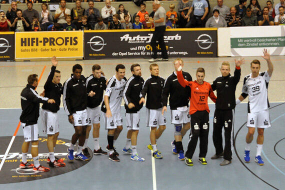 Eine Männerhandballmannschaft steht in einer Reihe auf einem Hallenplatz, einige Spieler winken der Menge zu. Sie tragen schwarze und weiße Trikots und Jacken. Die Zuschauer füllen die Tribünen hinter ihnen.