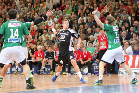 Ein Handballspieler in einem schwarzen Trikot versucht, während eines Spiels an zwei Verteidigern in grünen Trikots vorbeizukommen, während im Hintergrund eine Zuschauermenge von der Tribüne aus zusieht.