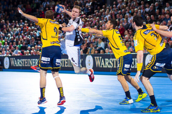 Ein Handballspieler in einem weiß-schwarzen Trikot springt, um den Ball zu werfen, während drei gegnerische Spieler in gelben Trikots versuchen, ihn während eines intensiven Spiels in einer überfüllten Arena zu blockieren.