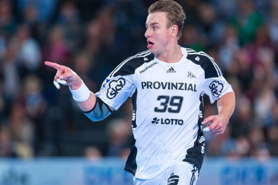 Ein männlicher Handballspieler in einem weiß-schwarzen Trikot mit der Nummer 39 läuft während eines Spiels und zeigt nach links, während im Hintergrund eine unscharfe Menschenmenge zu sehen ist.