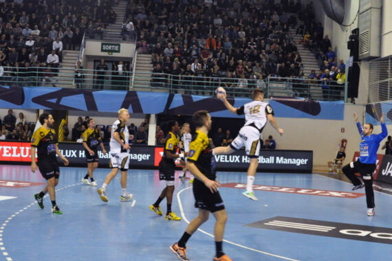 Ein Handballspieler in einem weißen Trikot springt zum Torschuss, während ein blau gekleideter Torwart sich darauf vorbereitet, den Ball zu blocken. Andere Spieler und ein großes Publikum verfolgen das Geschehen in einer Halle.