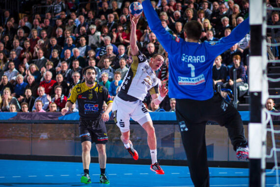 Ein Handballspieler in einem weiß-schwarzen Trikot springt und schießt auf das Tor, während der Torwart in Blau bereit steht, um das Tor zu verhindern. Die Zuschauer auf der Tribüne im Hintergrund schauen aufmerksam zu.