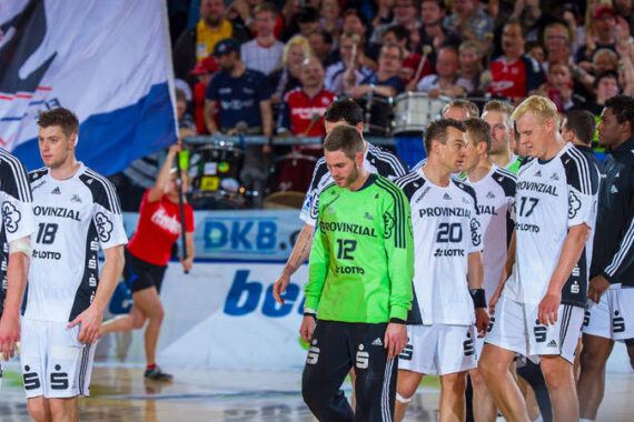 Eine Gruppe von männlichen Handballspielern in weißen und schwarzen Trikots geht auf ein Spielfeld, einer trägt ein grünes Torwarttrikot. Zuschauer mit Fahnen und Bannern füllen die Tribünen im Hintergrund.