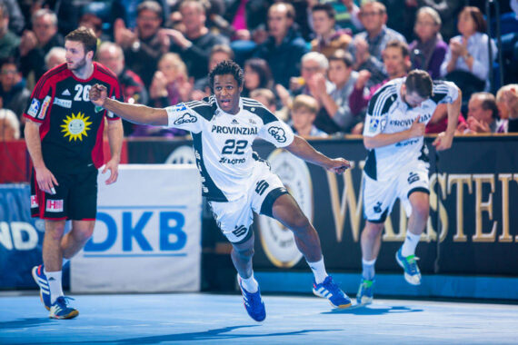 Ein Handballspieler in einem weißen Trikot mit der Nummer 22 jubelt mit ausgestreckten Armen auf einem blauen Spielfeld, während ein anderer Spieler hinterherläuft und ein Spieler in einem schwarzen Trikot zuschaut. Die Zuschauer schauen von der Tribüne im Hintergrund zu.