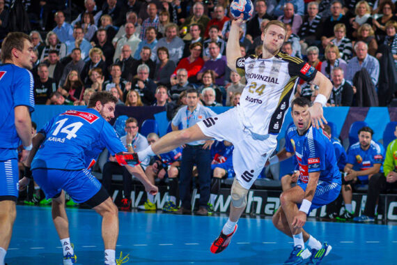Ein Handballspieler in einem weißen Trikot springt, um einen Wurf zu machen, während drei Spieler in blauen Trikots ihn verteidigen. Das Spiel findet auf einem Hallenplatz statt, im Hintergrund eine Zuschauermenge.