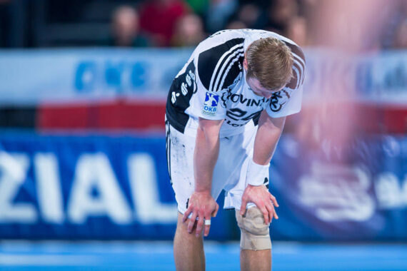 Ein Handballspieler in einem weiß-schwarzen Trikot beugt sich erschöpft mit den Händen auf den Knien über ein Hallenfeld mit einem verschwommenen Hintergrund von Zuschauern.