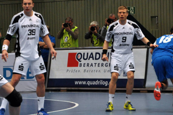 Zwei Handballspieler in weißen Trikots mit den Nummern 39 und 9 verteidigen gegen einen Gegner in Blau, während Fotografen in gelben Westen das Geschehen vom Spielfeldrand aus festhalten. Die Kulisse einer Sporthalle ist zu sehen.