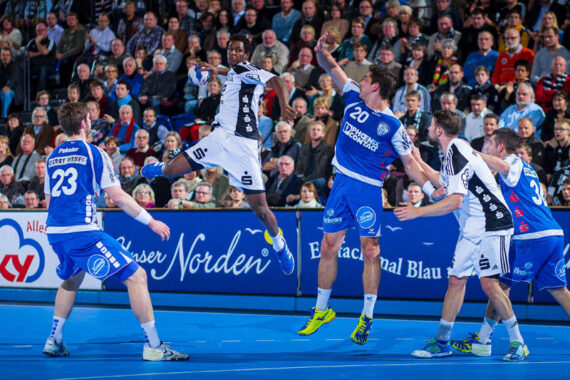 Ein Handballspieler in einem schwarz-weißen Trikot springt, um den Ball zu schießen, während Verteidiger in blauen Uniformen versuchen, ihn auf einem Hallenplatz zu blockieren, während im Hintergrund eine Menschenmenge zusieht.