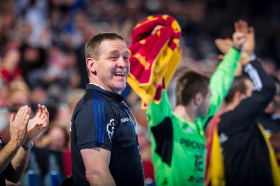 Ein lächelnder Mann in einem dunklen Sporthemd steht während einer Hallensportveranstaltung vor jubelnden Mannschaftskameraden, von denen einer ein rot-gelbes Handtuch schwenkt. Die Menge ist im Hintergrund verschwommen.