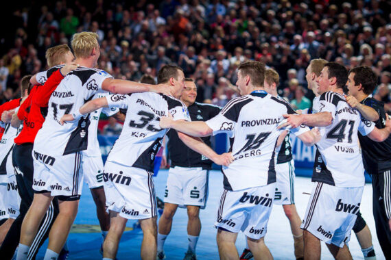 Eine Gruppe von männlichen Handballspielern in weißen Trikots feiert im Kreis auf einem Hallenplatz, lächelnd und sich in den Armen liegend, mit einem großen Publikum im Hintergrund.