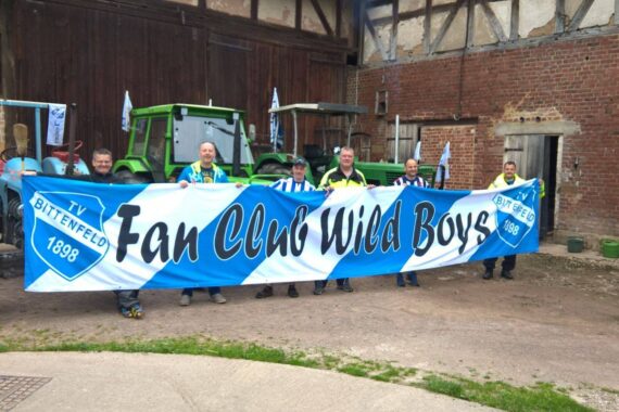 Sechs Personen stehen im Freien vor Traktoren und einem rustikalen Backsteingebäude und halten ein großes blau-weißes Transparent mit der Aufschrift Fan Club Wild Boys. Sie tragen Fankleidung und lächeln für das Foto.