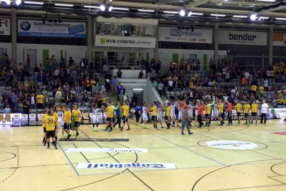Spieler in gelben und weißen Trikots geben sich auf einem Hallenhandballfeld die Hand, während die Schiedsrichter in der Nähe sind und eine große Menschenmenge von der Tribüne aus zusieht.