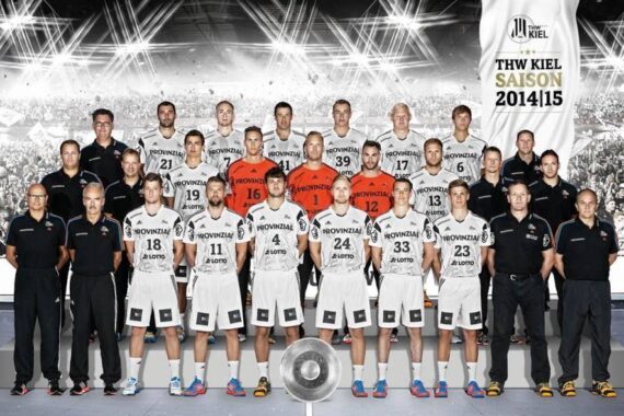Die Handballer des THW Kiel und ihre Betreuer posieren gemeinsam für ein Gruppenfoto in weiß-schwarzen Trikots vor einer Stadionkulisse und einem Transparent mit der Aufschrift THW Kiel Saison 2014/15 im Hintergrund.