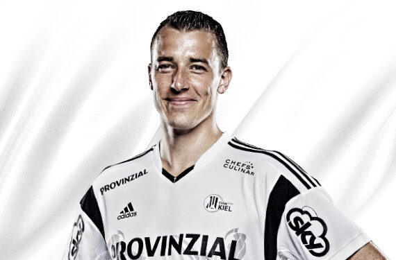 Ein männlicher Sportler in einem weißen Sporttrikot mit schwarzen Akzenten und mehreren Sponsorenlogos steht lächelnd mit den Händen in den Hüften vor einem weißen Hintergrund.