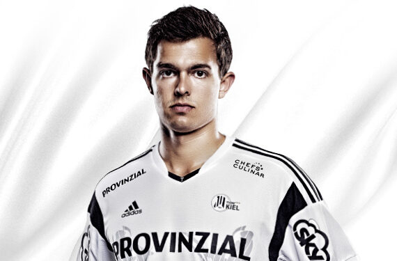 Ein junger Mann, der ein weißes Sporttrikot mit schwarzen Streifen und mehreren Sponsorenlogos trägt, steht vor einem weißen Hintergrund und schaut mit neutralem Blick direkt in die Kamera.
