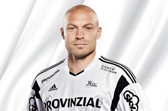 Ein Mann mit Glatze, der ein weißes und schwarzes Sporttrikot mit Sponsorenlogos trägt, steht vor einem weißen Hintergrund.