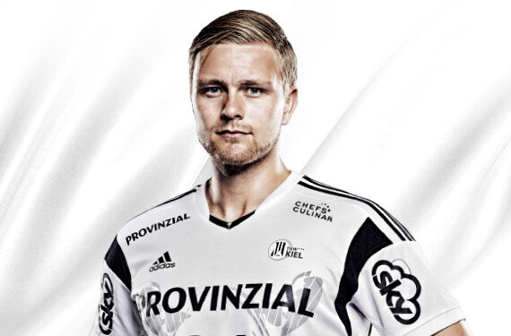 Ein männlicher Sportler in einem weiß-schwarzen Sporttrikot steht vor einem weißen Hintergrund und schaut mit neutralem Blick direkt in die Kamera. Auf dem Trikot sind mehrere Sponsorenlogos und ein Adidas-Logo zu sehen.
