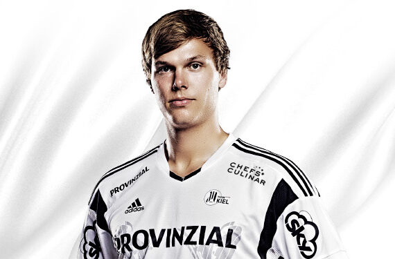 Ein junger männlicher Sportler mit kurzen braunen Haaren, der ein weißes Sporttrikot mit schwarzen Akzenten und verschiedenen Sponsorenlogos trägt, steht vor einem weißen Hintergrund.