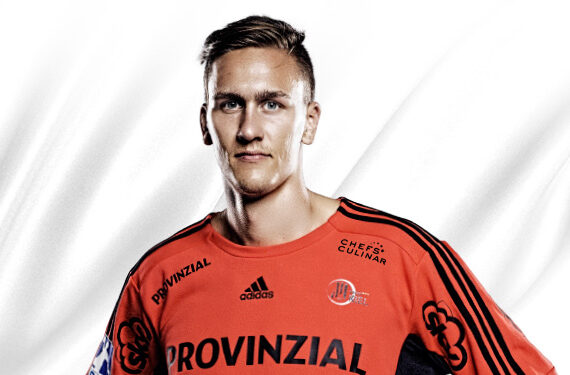 Ein Mann, der ein orangefarbenes Sporttrikot mit schwarzen Streifen und verschiedenen Sponsorenlogos trägt, steht vor einem weißen Hintergrund und blickt mit neutraler Miene in die Kamera.