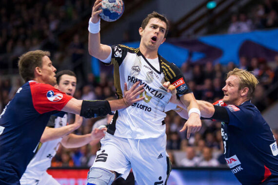Ein Handballspieler in einem weiß-schwarzen Trikot springt zum Wurf und hält den Ball über seinen Kopf, während zwei gegnerische Spieler in blauen und roten Trikots versuchen, ihn während eines Spiels zu blockieren. Im Hintergrund sind Zuschauer zu sehen.