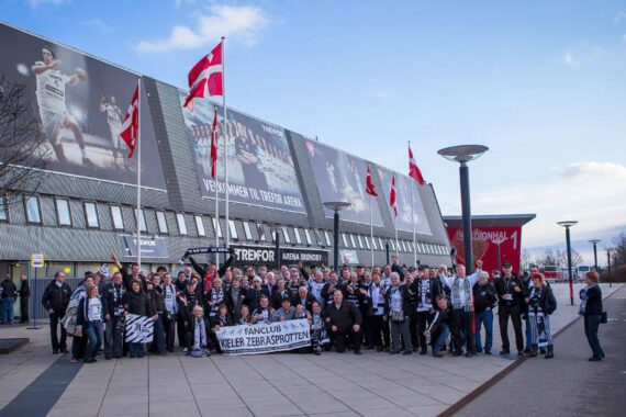 Eine große Gruppe von Sportfans mit Schals und Fahnen posiert vor einer Arena und hält ein Transparent mit der Aufschrift Fanclub Keler ZeBarspiotten. Über der Gruppe sind dänische Fahnen zu sehen.