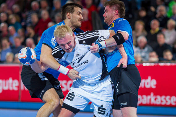 Zwei Spieler in blauen Trikots verteidigen sich während eines intensiven Spiels gegen einen Spieler in einem weißen Trikot, der einen Handball hält, während die Zuschauer im Hintergrund zuschauen.